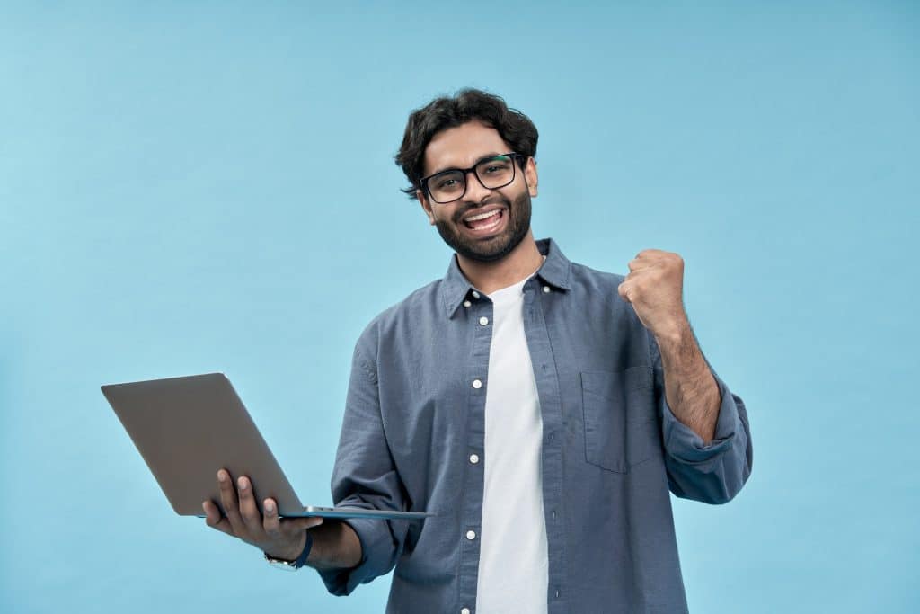 Happy young arab man celebrating win holding laptop isolated on blue.