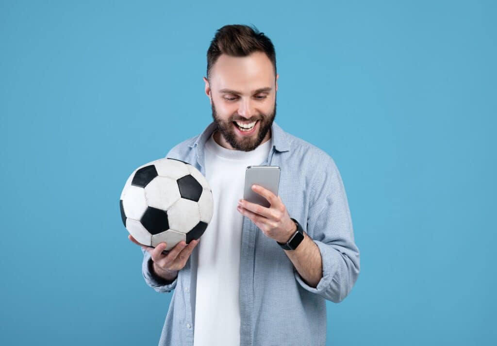 Happy young guy with soccer ball using smartphone, winning sports bet, rooting for his favorite team