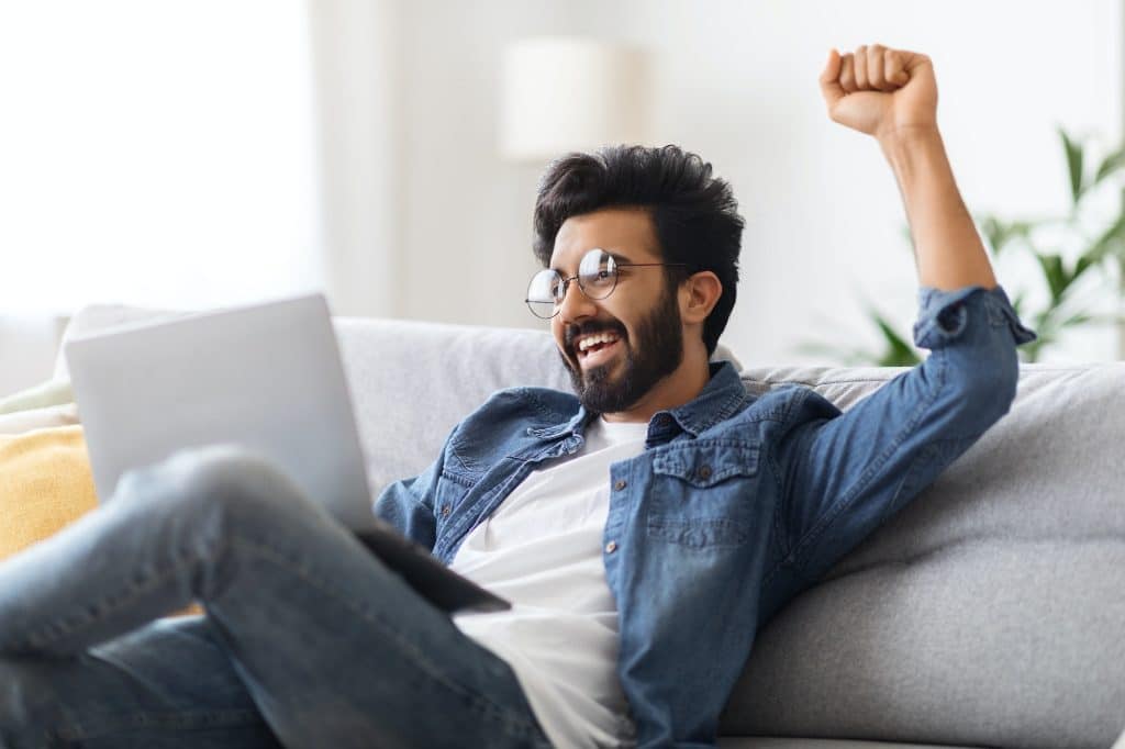 Online Win. Overjoyed Young Indian Man Celebrating Success With Laptop At Home