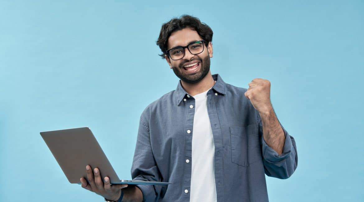 Happy young arab man celebrating win holding laptop isolated on blue.